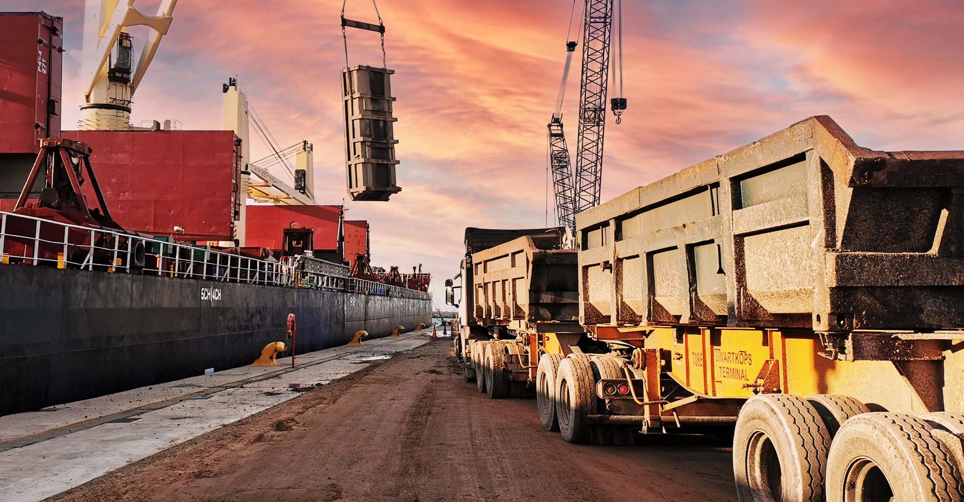 dump truck lined up on a dock beside a cargo ship, while a crane lifts heavy cargo