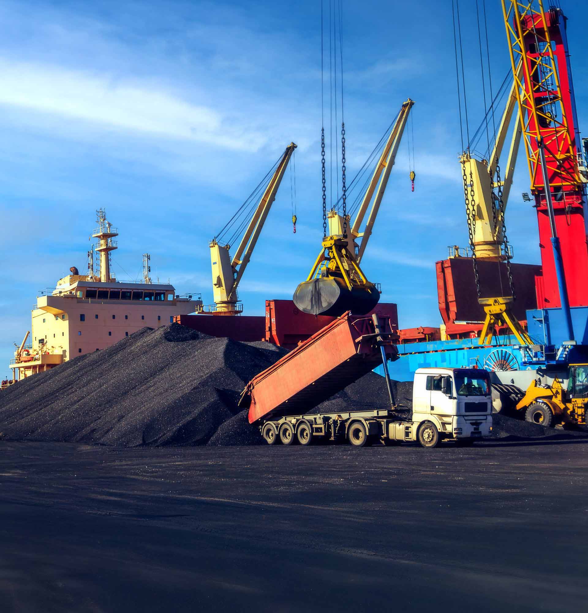 truck unloading coal onto a large pile at an industrial port, with cranes, heavy machinery and a cargo ship in the background