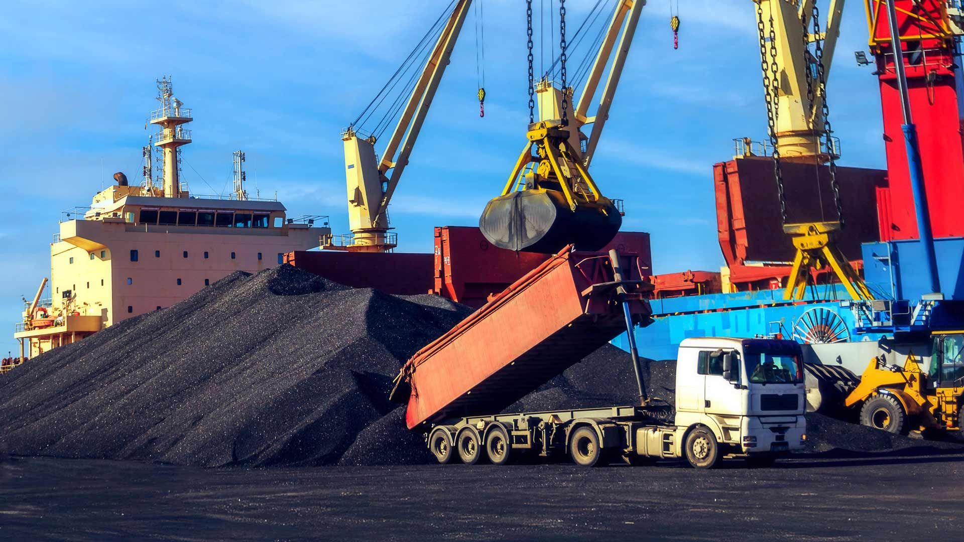 truck unloading coal onto a large pile at an industrial port, with cranes, heavy machinery and a cargo ship in the background